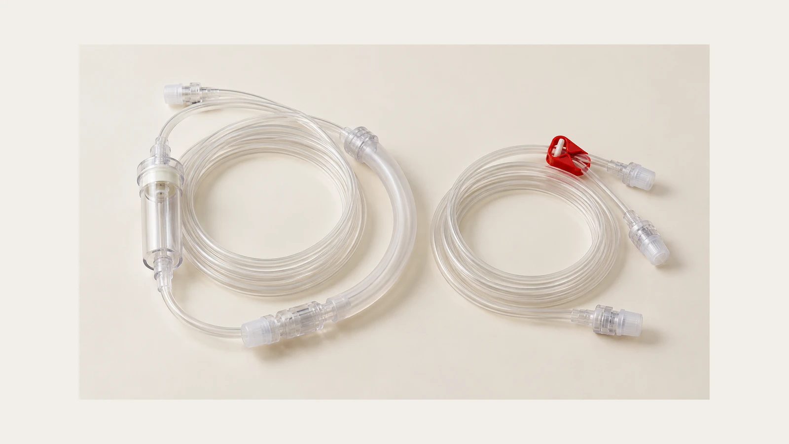 Illustrative still life of clear coiled bloodline components with capped connectors, an inline chamber and a red clamp on an off-white surface.