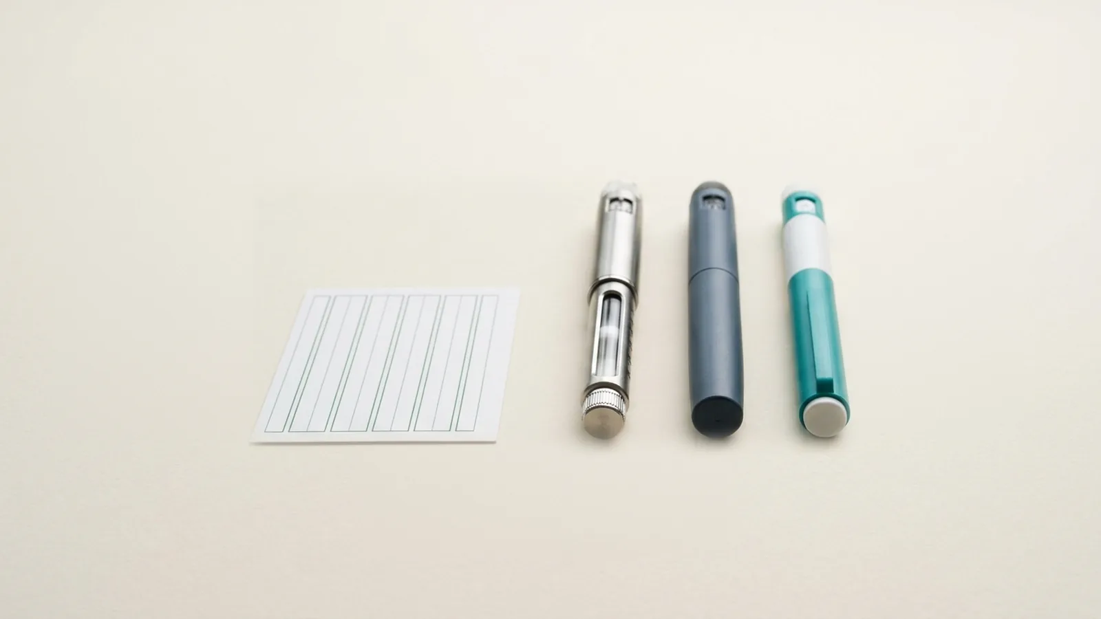 Three injection pens with different body styles, shown without needles attached, lying beside a blank card ruled with forest-green lines on a warm off-white surface